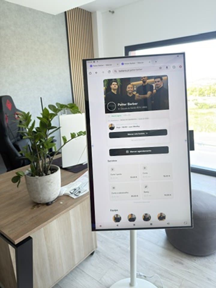 Totem BarberIA num espaço de atendimento, com o ecrã vertical a mostrar os serviços disponíveis e a equipa da barbearia.